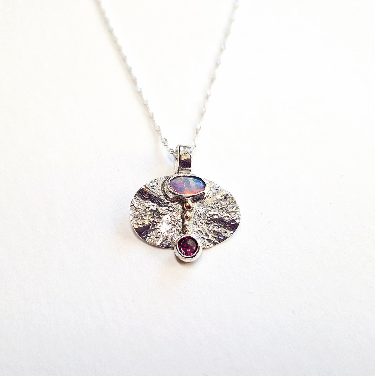 Silver necklace with a round pendant featuring an opal and a red gemstone on a white background