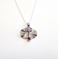Silver necklace with a round pendant featuring an opal and a red gemstone on a white background